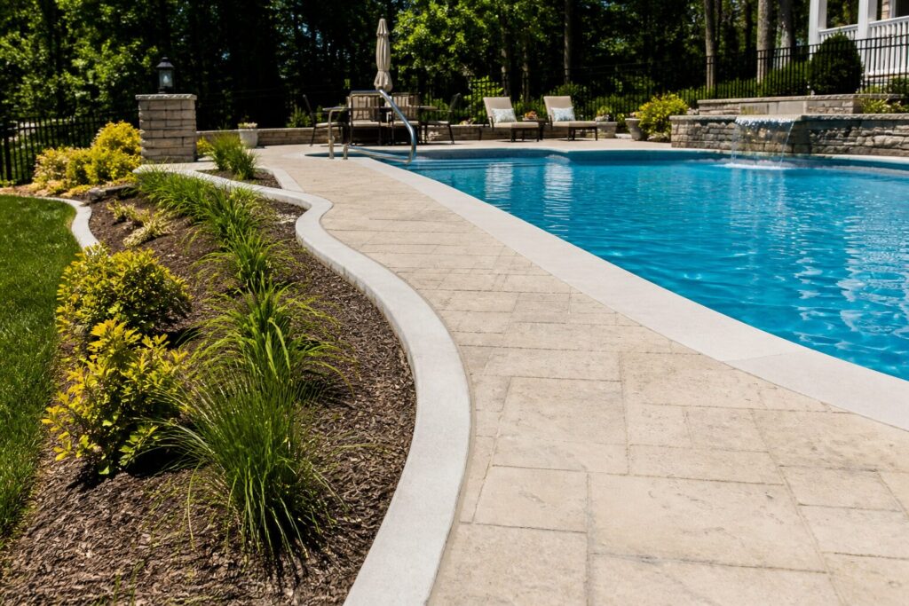 Curved concrete curbing borders a landscaped bed beside a stone pool deck in a sunny backyard